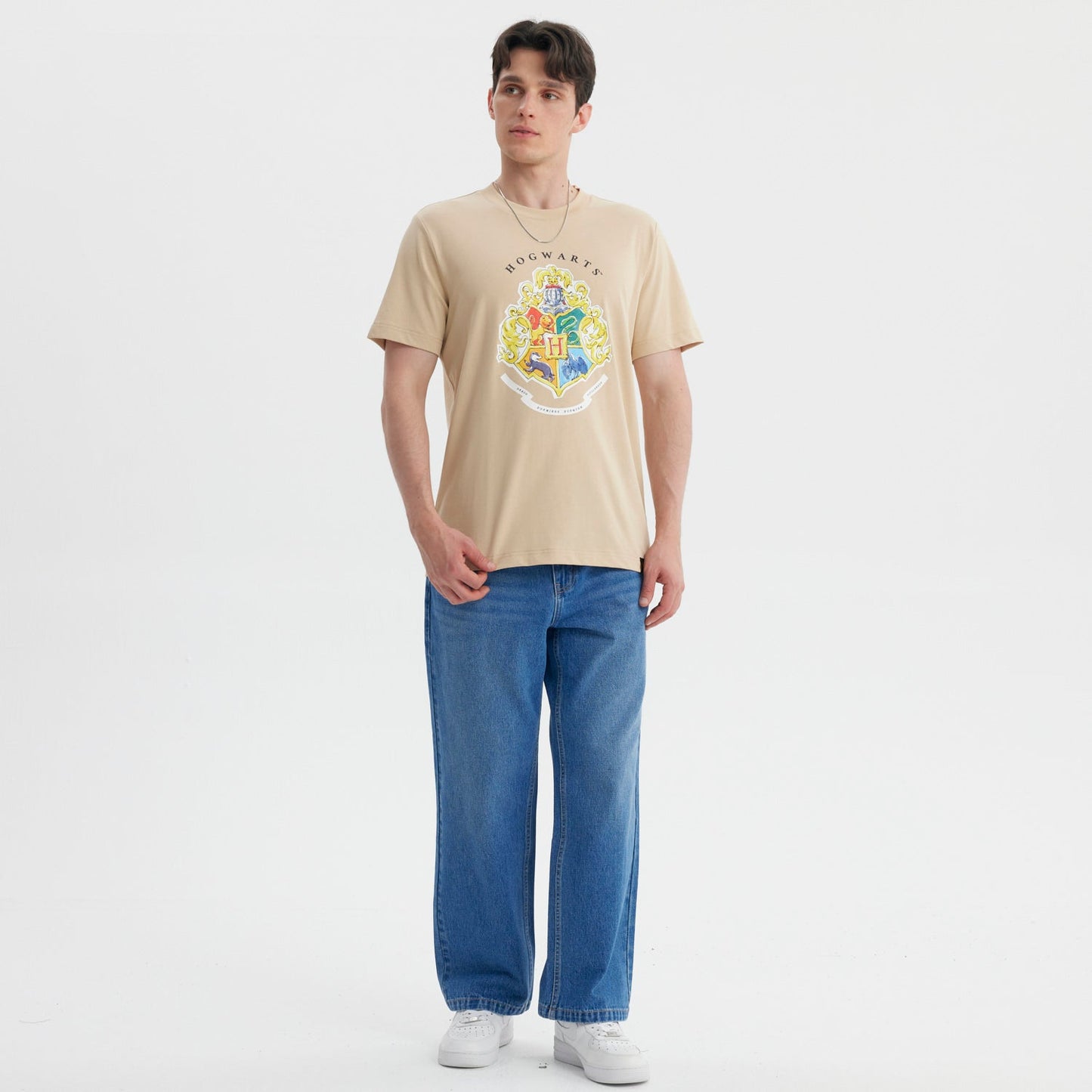 Polera beige de algodón con el escudo de Hogwarts en colores vibrantes, destacando los cuatro emblemas de las casas y la letra H, ideal para fans de Harry Potter.