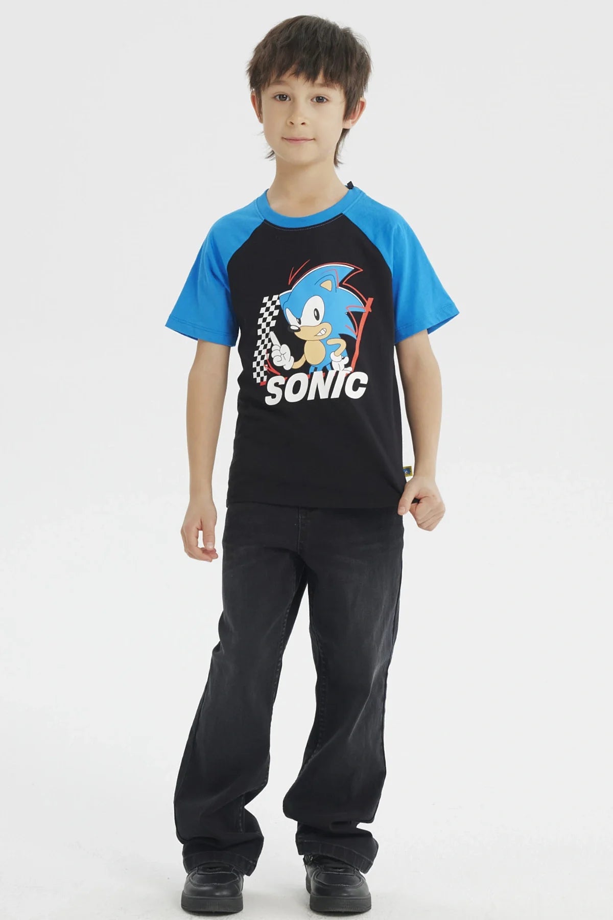 Polera negra con detalles en azul, presenta a Sonic en un estilo cartoon, con fondo de banderas a cuadros y el texto 'SONIC' en blanco, ideal para fans de videojuegos.